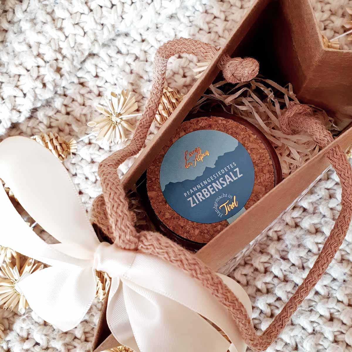 Detailaufnahme eines Tontöpfchens mit Tiroler Zirbensalz von Essenz der Alpen in einer rustikalen Geschenkschachtel mit dekorativen Bändern und Strohsternen.