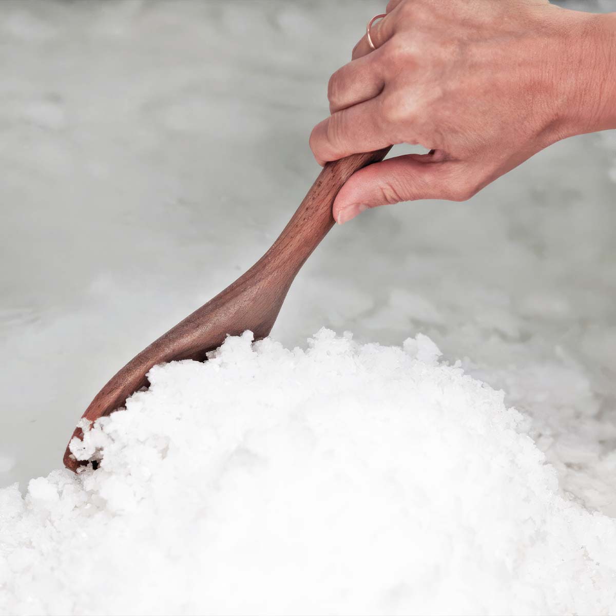 Nahaufnahme der traditionellen Salzgewinnung bei Essenz der Alpen: Eine Hand schiebt frisch gebildete weiße Salzkristalle mit einem Holzschaber zusammen.