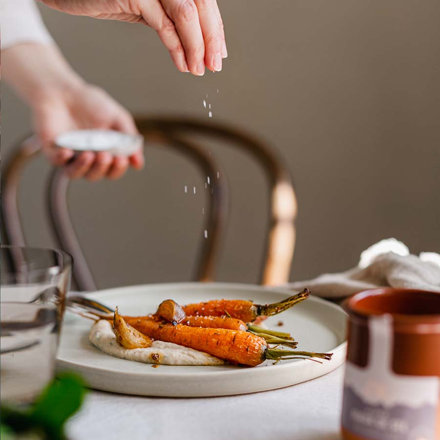 Eine Hand streut feine Salzkristalle über geröstete Karotten auf cremigem Püree. Daneben steht ein Tontöpfchen mit Alpen Fleur de Sel von Essenz der Alpen.