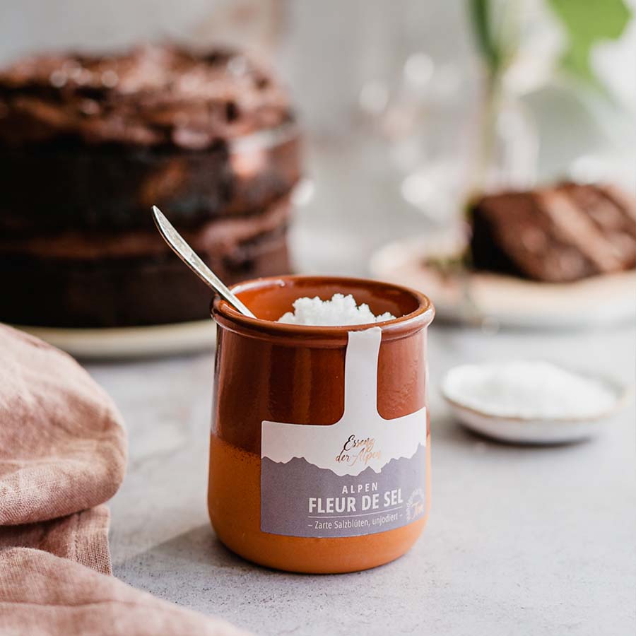 Geöffnetes Tontöpfchen mit handgeschöpftem Alpen Fleur de Sel von Essenz der Alpen und einem kleinen Löffel, dekorativ angerichtet neben einem Kuchen.