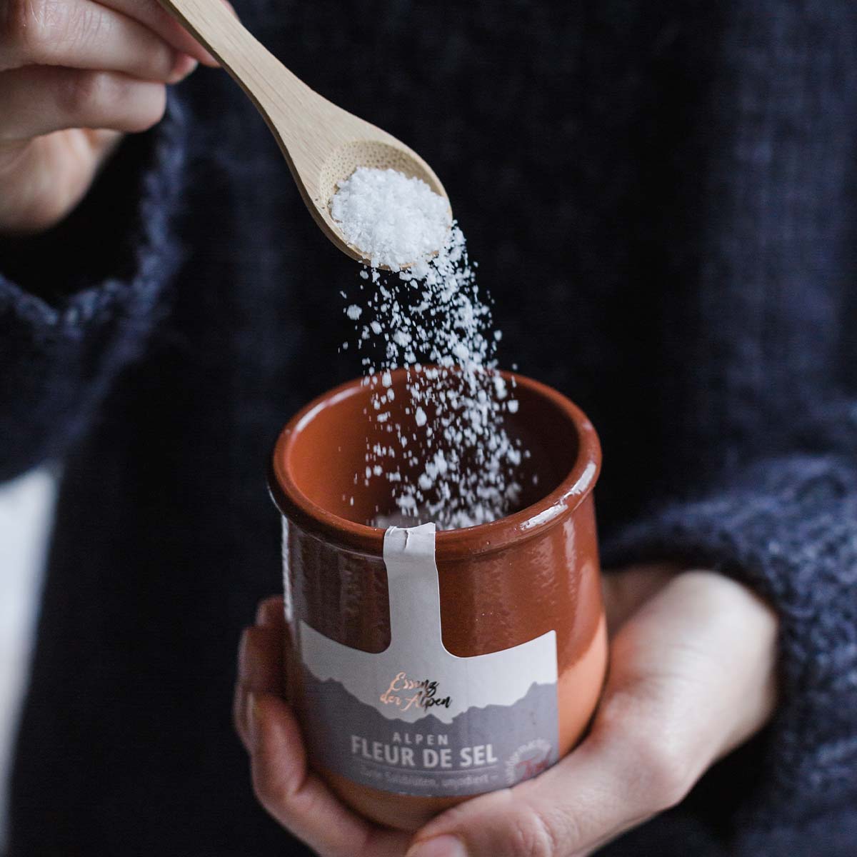 Eine Mitarbeiterin füllt feine Salzkristalle mit einem Holzlöffel in ein braunes Tontöpfchen mit der Aufschrift Alpen Fleur de Sel von Essenz der Alpen.