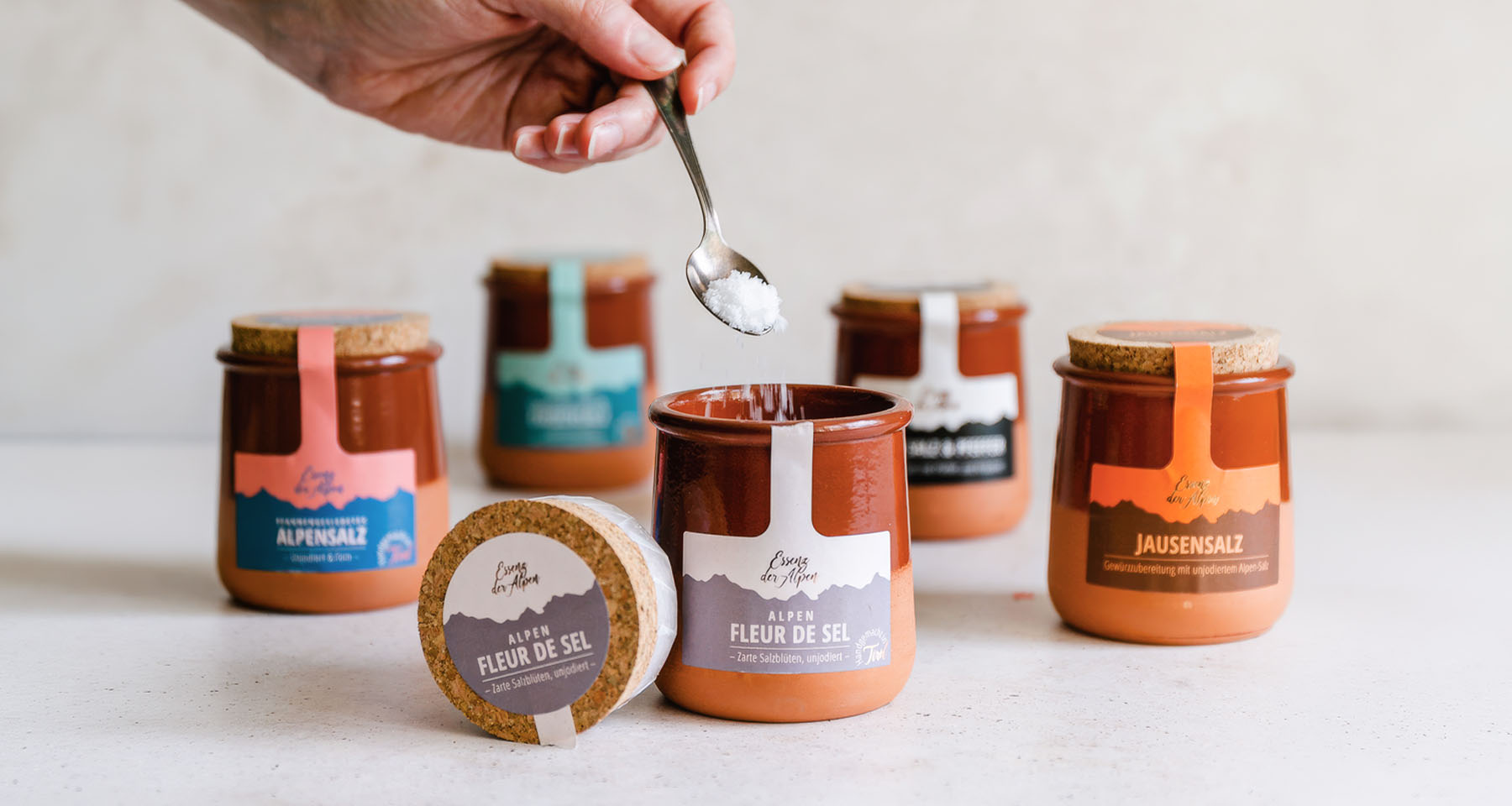 Alpen Fleur de Sel von Essenz der Alpen im geöffneten Tontöpfchen, während eine Hand Salzflocken mit einem Löffel hineinstreut. Weitere Tiroler Salzspezialitäten stehen im Hintergrund.