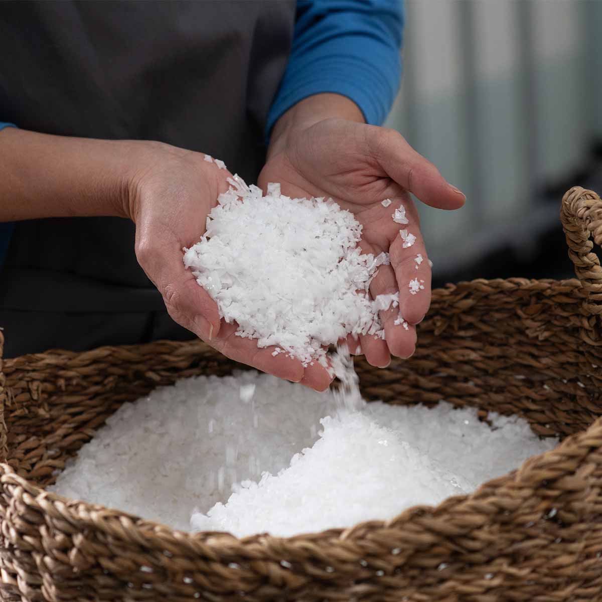 Zwei Hände halten frisch geschöpfte, weiße Salzflocken über einem geflochtenen Korb mit Salz. Nahaufnahme der handwerklichen Salzverarbeitung.