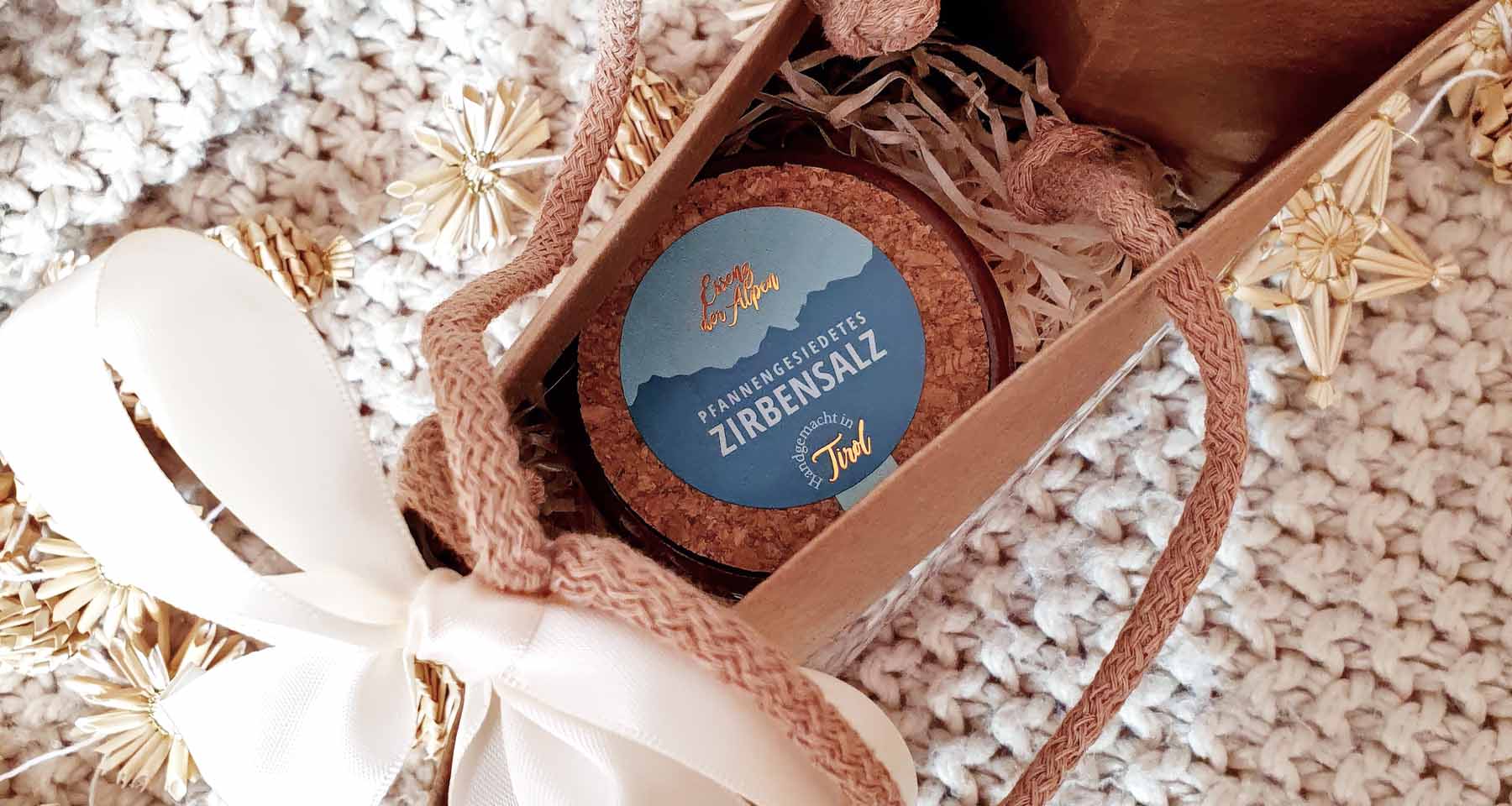 Pfannengesiedetes Zirbensalz von Essenz der Alpen in einer dekorativen Geschenkschachtel mit Korkdeckel, Schleifen und Strohsternen auf einer gestrickten Decke.